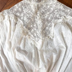 May include: Close-up of a white vintage blouse with intricate lace detailing. The high collar and yoke feature delicate floral patterns, while the body of the blouse has textured panels. The fabric appears soft and lightweight, suggesting a delicate garment.
