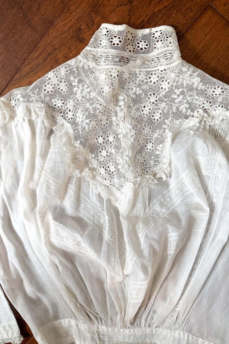 May include: Close-up of a white vintage blouse with intricate lace detailing. The high collar and yoke feature delicate floral patterns, while the body of the blouse has textured panels. The fabric appears soft and lightweight, suggesting a delicate garment.