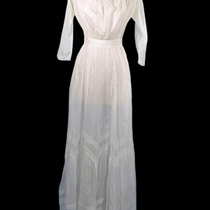 May include: A vintage white dress with long sleeves and a high neckline. The dress features delicate lace detailing around the neck and a fitted waist, flowing into a full skirt. The dress is displayed on a mannequin against a black background.