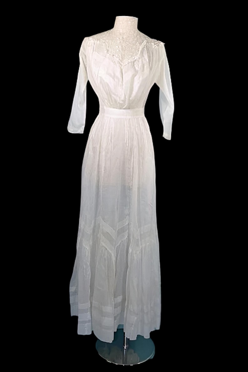 May include: A vintage white dress with long sleeves and a high neckline. The dress features delicate lace detailing around the neck and a fitted waist, flowing into a full skirt. The dress is displayed on a mannequin against a black background.