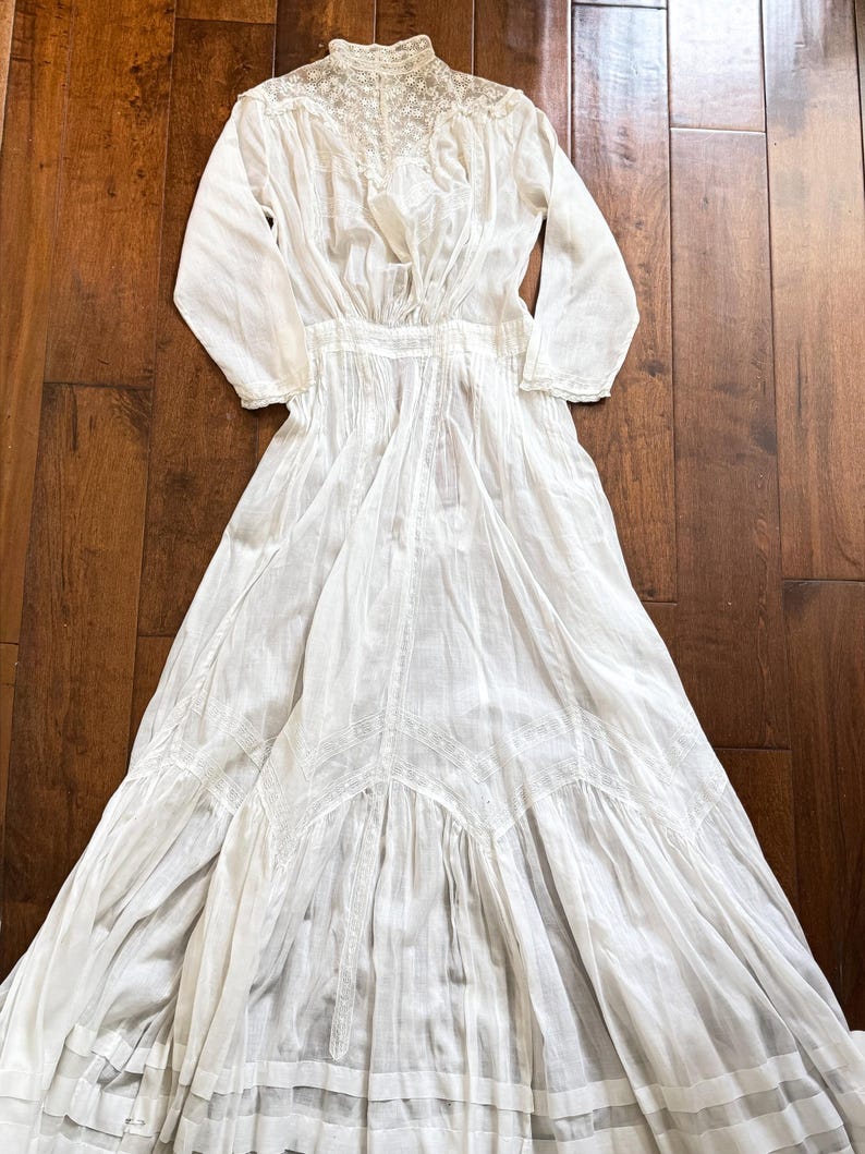 May include: A white vintage dress with long sleeves, a high neckline, and intricate lace detailing. The dress features a fitted bodice and a flowing skirt, showcasing a classic, elegant design. The dress is made of a lightweight fabric.
