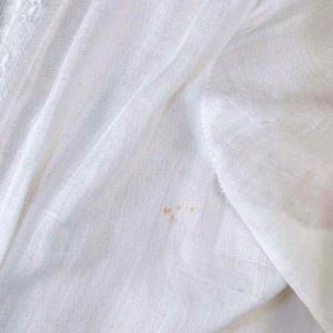 May include: Close-up of a white fabric with a textured weave. The fabric has a soft, flowing appearance with subtle folds and creases. Small, light brown stains are visible on the fabric's surface, adding a touch of character.