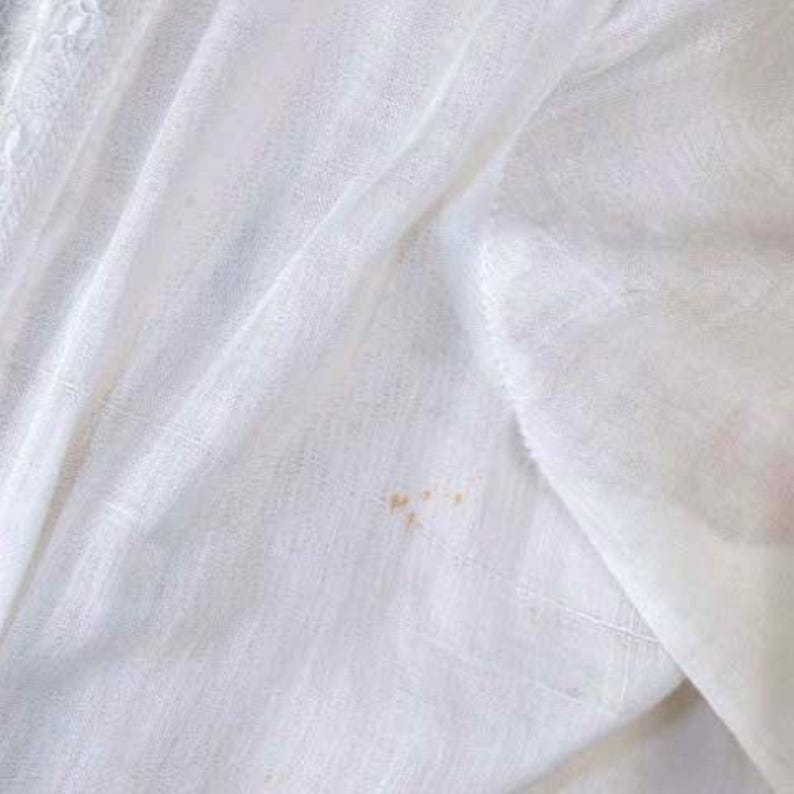 May include: Close-up of a white fabric with a textured weave. The fabric has a soft, flowing appearance with subtle folds and creases. Small, light brown stains are visible on the fabric's surface, adding a touch of character.