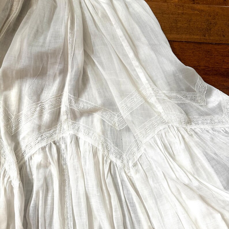 May include: A white, flowing skirt with intricate lace detailing. The fabric appears lightweight and airy, with a delicate pattern along the hem and seams. The skirt is gathered, creating soft folds and a romantic aesthetic.