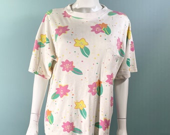Puede incluir: Camiseta blanca de manga corta con estampado floral en rosa, amarillo y verde. La camiseta tiene un pequeño bolsillo en el lado izquierdo.