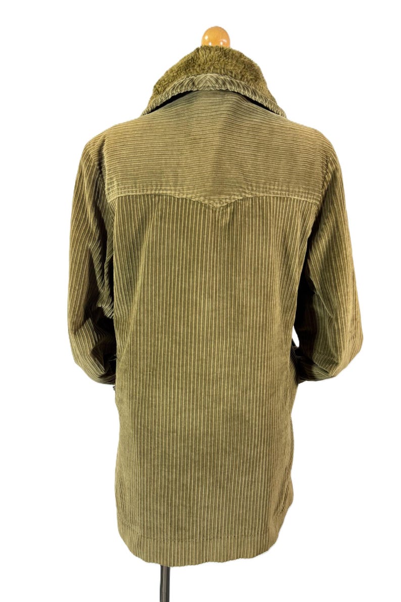 May include: Olive green corduroy jacket with a faux fur collar. The jacket has a button-up front and long sleeves. The back of the jacket is visible, showing vertical corduroy lines and a yoke detail. The jacket is displayed on a hanger.