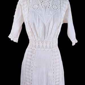 May include: A white vintage dress with intricate lace detailing. The dress features a high neckline, short sleeves, and a fitted waist. The bodice and sleeves have a ribbed texture, and the skirt is long and flowing. The dress is likely made of cotton or linen.