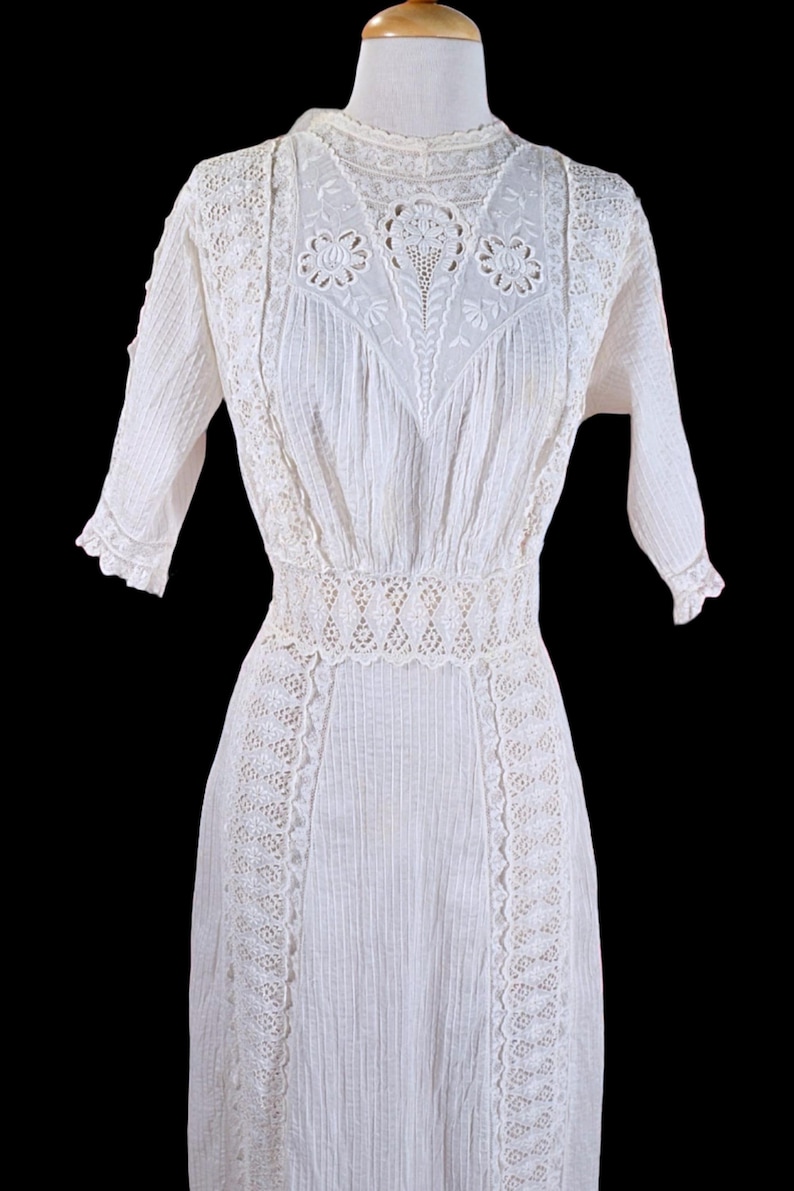 May include: A white vintage dress with intricate lace detailing. The dress features a high neckline, short sleeves, and a fitted waist. The bodice and sleeves have a ribbed texture, and the skirt is long and flowing. The dress is likely made of cotton or linen.