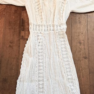 May include: A white vintage dress with long sleeves, featuring intricate lace detailing. The dress has a high neckline and a fitted waist, with vertical pleats on the skirt. The lace is used on the sleeves, bodice, and skirt.