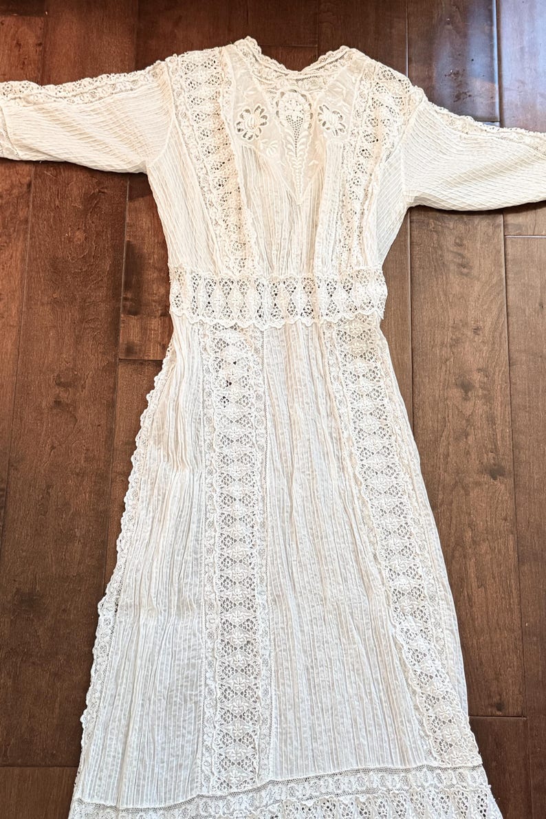 May include: A white vintage dress with long sleeves, featuring intricate lace detailing. The dress has a high neckline and a fitted waist, with vertical pleats on the skirt. The lace is used on the sleeves, bodice, and skirt.