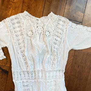 May include: A white vintage dress with intricate lace detailing. The dress features a high neckline, short sleeves, and a fitted bodice. The lace is used on the sleeves, neckline, and bodice. The dress is made of a lightweight fabric, perfect for a summer day.