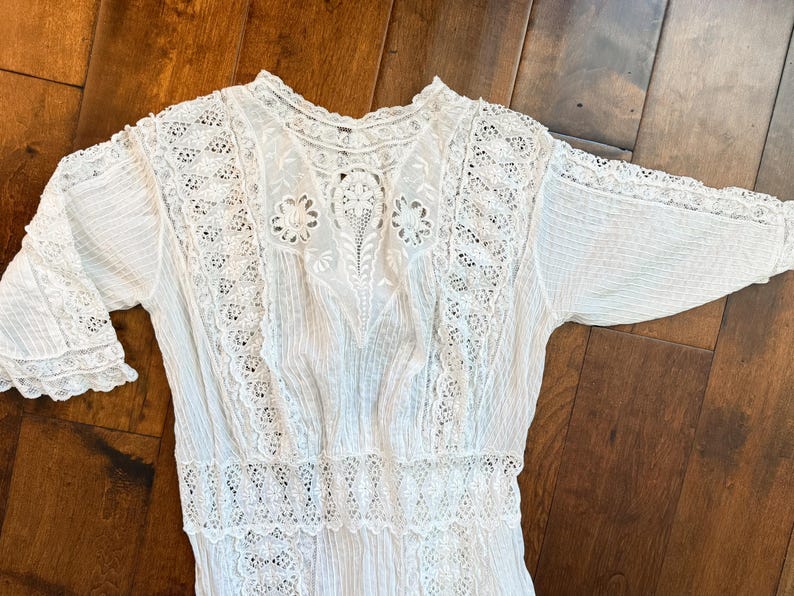 May include: A white vintage dress with intricate lace detailing. The dress features a high neckline, short sleeves, and a fitted bodice. The lace is used on the sleeves, neckline, and bodice. The dress is made of a lightweight fabric, perfect for a summer day.