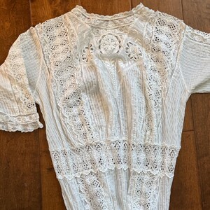 May include: A white vintage blouse with intricate lace detailing. The blouse features a high neckline, short sleeves, and a fitted waist. The lace is used on the sleeves, bodice, and hem, creating a delicate and elegant look.