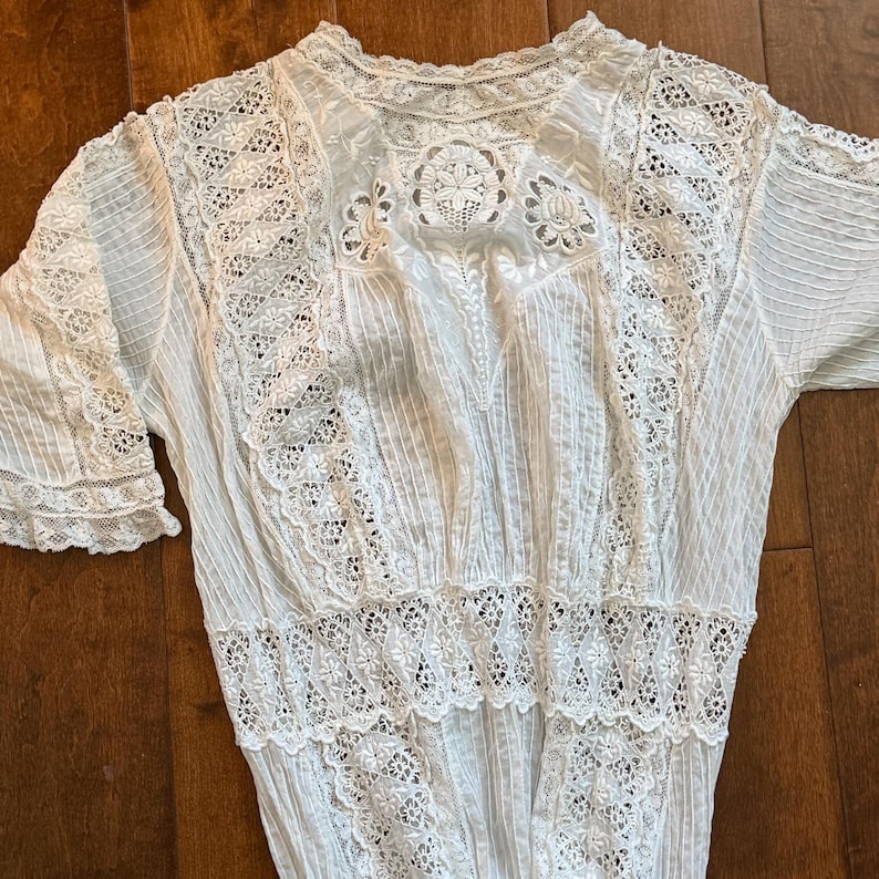 May include: A white vintage blouse with intricate lace detailing. The blouse features a high neckline, short sleeves, and a fitted waist. The lace is used on the sleeves, bodice, and hem, creating a delicate and elegant look.