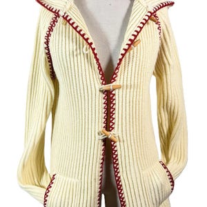 May include: Cream-colored hooded cardigan with red trim and wooden toggle closures. The ribbed knit sweater features two front pockets and a hood. The edges are accented with a decorative red stitch.