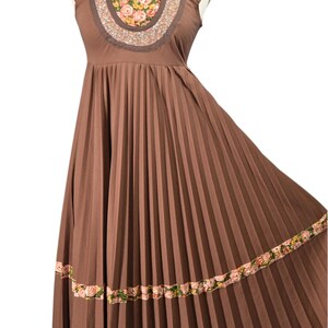 May include: Brown pleated dress with a floral embroidered yoke and hemline. The dress features a square neckline and cap sleeves. The floral embroidery includes pink, yellow, and green accents. The dress is displayed on a mannequin.