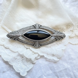 Puede incluir: Un broche de plata con una piedra de ónix negro engastada en un diseño geométrico. El broche está rodeado de piedras de marcasita.