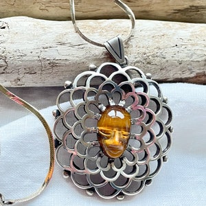 Op de afbeelding: Zilveren ketting met een hanger met een tijgeroog-steen in de vorm van een gezicht. De hanger is omgeven door een delicaat, cirkelvormig ontwerp.