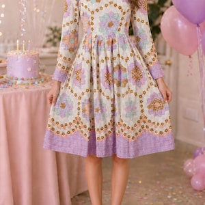 May include: A long-sleeved dress featuring a cream base with a repeating floral and geometric pattern in shades of lavender, light blue, and brown. The dress has a round neckline, a fitted bodice, and a pleated skirt that falls to the knees.