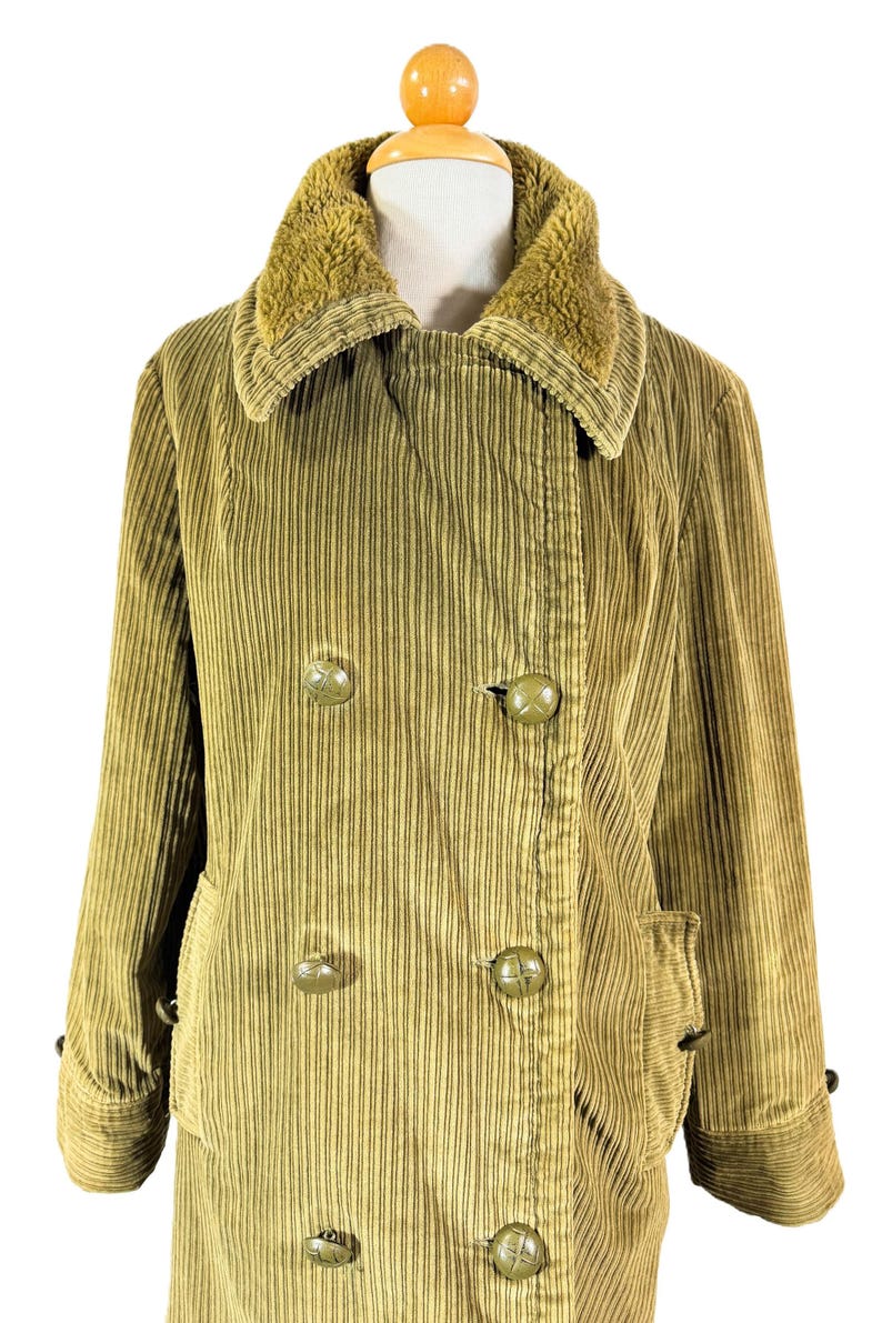 May include: A corduroy coat in a muted olive green color, featuring a double-breasted design with large, round buttons. The coat has a plush, textured collar and side pockets. The garment is displayed on a mannequin.