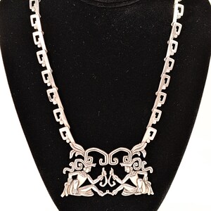 May include: A silver necklace with a pendant featuring two stylized figures with headdresses. The chain is made of square links.