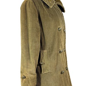 May include: A corduroy coat in a muted olive green color. The coat features a plush, textured collar and a double-breasted design with large, round buttons. The sleeves have buttoned cuffs, and there is a pocket on the front.