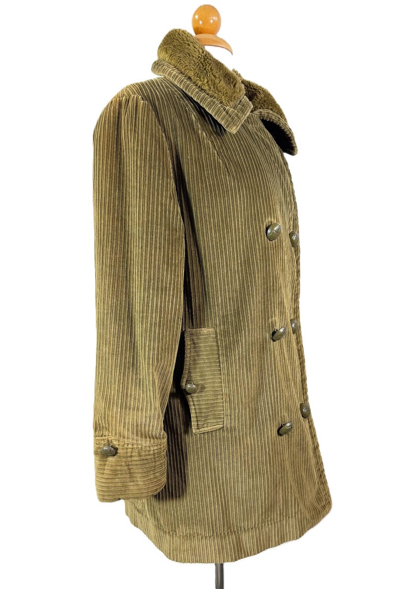 May include: A corduroy coat in a muted olive green color. The coat features a plush, textured collar and a double-breasted design with large, round buttons. The sleeves have buttoned cuffs, and there is a pocket on the front.