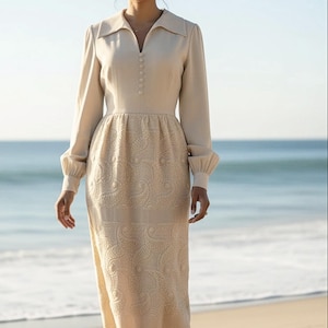 May include: A long, cream-colored dress with a collared neckline and long sleeves. The dress features a series of buttons down the front and an embroidered pattern on the skirt. The dress is worn on a beach with the ocean in the background.