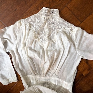 May include: A vintage white blouse with long sleeves. The blouse features a high, lace-trimmed collar and delicate floral embroidery on the yoke. The fabric appears lightweight and semi-sheer, with a gathered waist and subtle detailing.