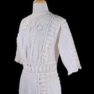 May include: A white vintage dress with intricate lace and embroidery details. The dress features a high neckline, short sleeves, and a fitted waist. The bodice and sleeves are adorned with delicate lace trim, and the skirt has vertical pleats.
