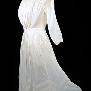 May include: A vintage white dress with long sleeves and a high, lace-trimmed neckline. The dress features a fitted waist and a flowing skirt with decorative lace panels near the hem. The fabric appears lightweight and semi-sheer.