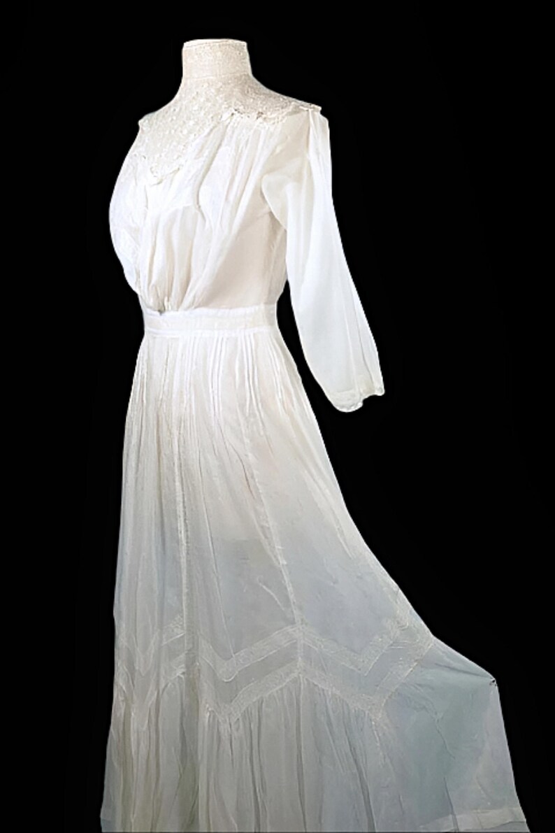 May include: A vintage white dress with long sleeves and a high, lace-trimmed neckline. The dress features a fitted waist and a flowing skirt with decorative lace panels near the hem. The fabric appears lightweight and semi-sheer.