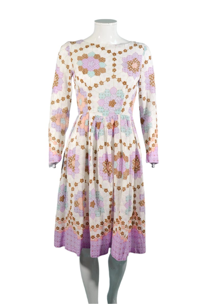 May include: A white dress with a long sleeve and a floral print. The print features a patchwork design with purple, pink, brown, and blue flowers. The dress has a fitted bodice and a full skirt.