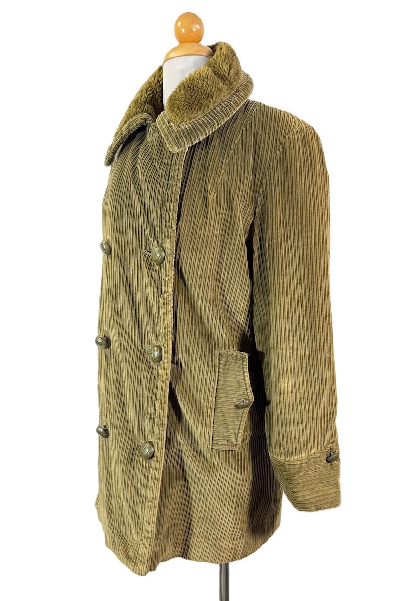 May include: A corduroy coat in olive green, displayed on a mannequin. The coat features a double-breasted design with large, round buttons and a plush, brown faux-fur collar. It has a pocket on the front and buttoned cuffs.
