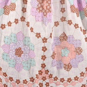 May include: White fabric with a repeating pattern of brown, pink, and purple flowers. The flowers are made up of smaller patchwork pieces. The fabric has a light, airy feel.