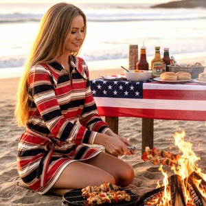 May include: A beach scene with a woman grilling kebabs over a small charcoal grill. She is wearing a red, white, and navy striped dress. An American flag serves as a tablecloth for a picnic table with condiments and buns.