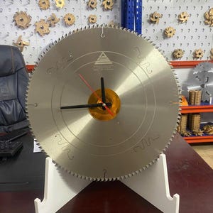 Puede incluir: Un disco de sierra circular reutilizado como reloj. La hoja plateada tiene un borde dentado y está montada sobre un soporte blanco. Las manecillas del reloj son negras, con un segundero rojo, y el centro es dorado.
