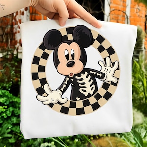May include: A white t-shirt featuring a graphic of a cartoon mouse character wearing a black skeleton costume. The character is positioned inside a circular frame with a black and beige checkered pattern, with both hands reaching outward.