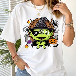 Puede incluir: Una camiseta blanca con un gráfico de un alienígena verde de tres ojos que lleva un sombrero de bruja negro con patrón de telaraña, un parche negro con una calavera y una capa negra, junto a una calabaza, dulces y una poción.