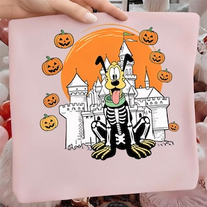 Op de afbeelding: Een lichtroze t-shirt met een grafische print van een cartoonhond in een skeletkostuum, zittend voor het silhouet van een wit kasteel tegen een grote oranje maan, omringd door verschillende kleine oranje pompoenen met gezichten.