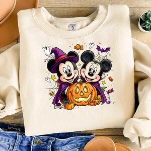 May include: A cream-colored crewneck sweatshirt featuring a graphic of two cartoon mice dressed in purple witch and vampire costumes. They are holding a large orange jack-o'-lantern surrounded by small ghosts, spiderwebs, bats, and candy.