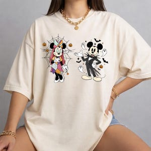 May include: An off-white short-sleeve t-shirt featuring a graphic print of two cartoon characters dressed in Halloween costumes. One character stands before a spiderweb with small pumpkins, while the other is surrounded by ghosts, bats, and pumpkins.