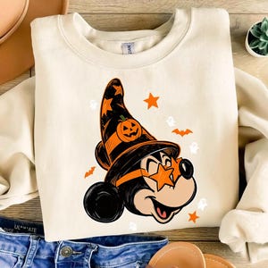 Puede incluir: Una sudadera de cuello redondo color crema con un gráfico de una cabeza de ratón de dibujos animados que lleva un sombrero de bruja negro y naranja con un diseño de calabaza. El ratón lleva gafas naranjas en forma de estrella, rodeado de pequeñas estrellas naranjas, murciélagos y fantasmas blancos.