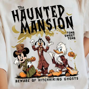 May include: A cream-colored t-shirt featuring a graphic of Mickey Mouse, Goofy, and Donald Duck dressed in spooky attire. The text reads The Haunted Mansion, Tomb Sweet Tomb, and Beware of Hitchhiking Ghosts, accompanied by small pumpkins and bats.
