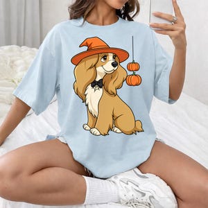 Op de afbeelding: Een lichtblauw t-shirt met een cartoonillustratie van een beige spaniël die een oranje heksenhoed en een zwarte vlinderdas draagt. De hond staat naast twee kleine oranje pompoenen die aan een dunne zwarte lijn op het shirt hangen.