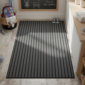 Op de afbeelding: Een rechthoekige deurmat met een grijs en zwart gestreept ontwerp. De mat heeft verticale zwarte strepen op een grijze achtergrond. Hij ligt op een betegelde vloer. Een mand met een rugzak en schoenen is zichtbaar op de achtergrond.