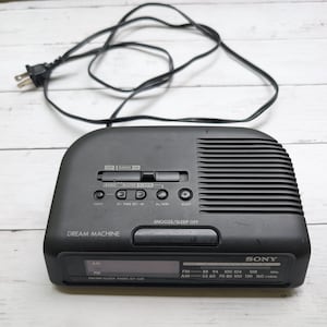 Puede incluir: Un radio reloj FM/AM Sony Dream Machine modelo ICF-C26 de color negro con pantalla digital, un botón de snooze/sleep off en la parte superior, varios interruptores de control para funciones de alarma y radio, y un cable de alimentación negro.
