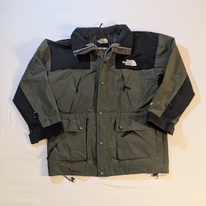 May include: A green and black The North Face jacket. The jacket has a black collar, shoulders, and sleeves. It features a button-up front, multiple pockets, and the brand's logo on the chest. The jacket is designed for outdoor wear.