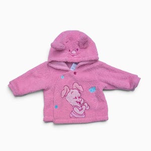 May include: A pink fleece hooded jacket featuring a textured surface, long sleeves, and a front button closure. The hood includes embroidered pig ears and facial features, while the front displays an embroidered graphic of a small pig character.