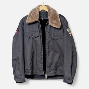 Puede incluir: Chaqueta azul marino oscuro con cuello de piel sintética y dos bolsillos en el pecho. La chaqueta tiene cierre de cremallera y parches decorativos en las mangas. La chaqueta está colgada de una percha de madera.